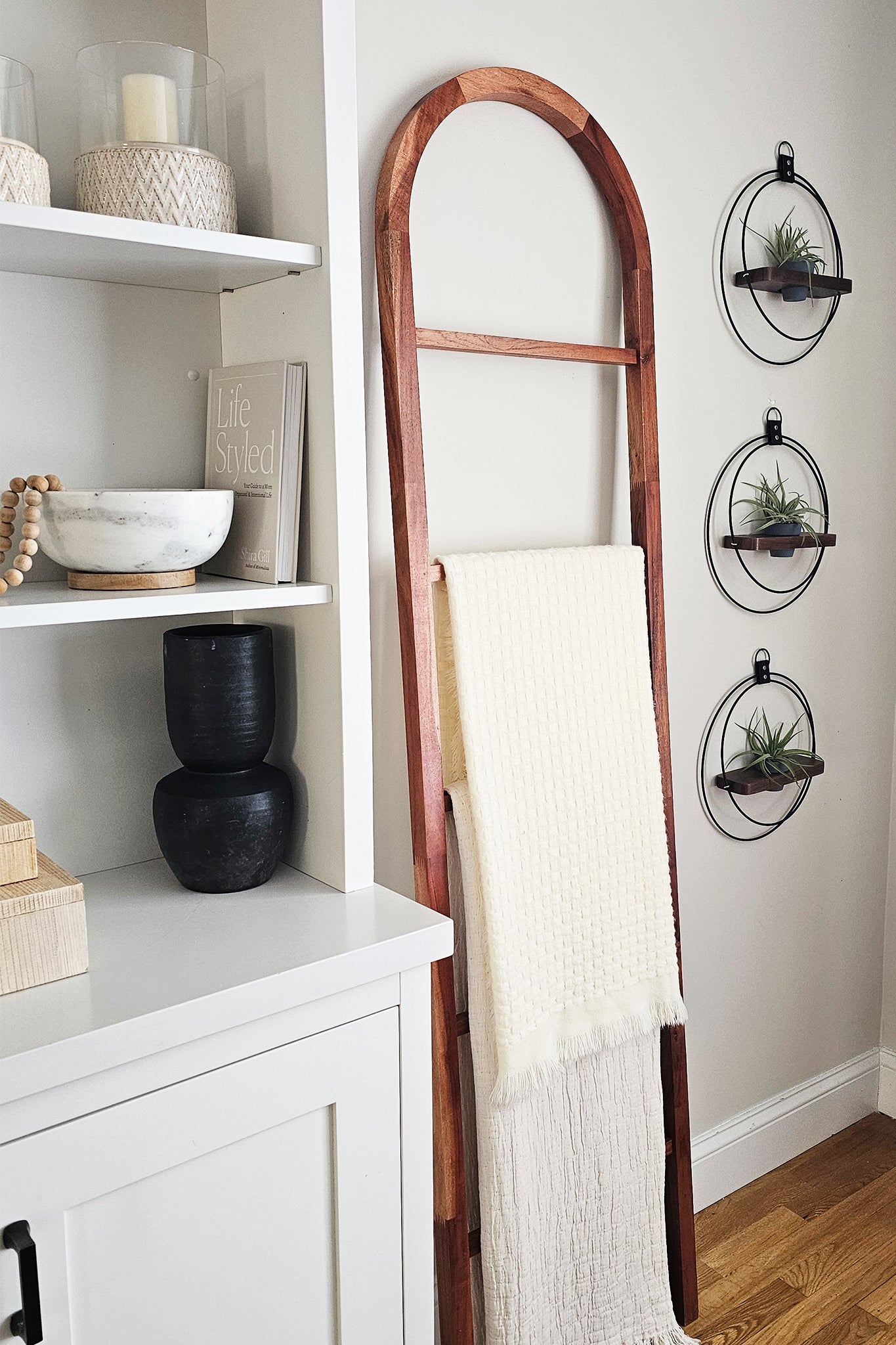 Wooden blanket ladder against a white wall with modern wall decor next to it featuring air plants and a small pot.